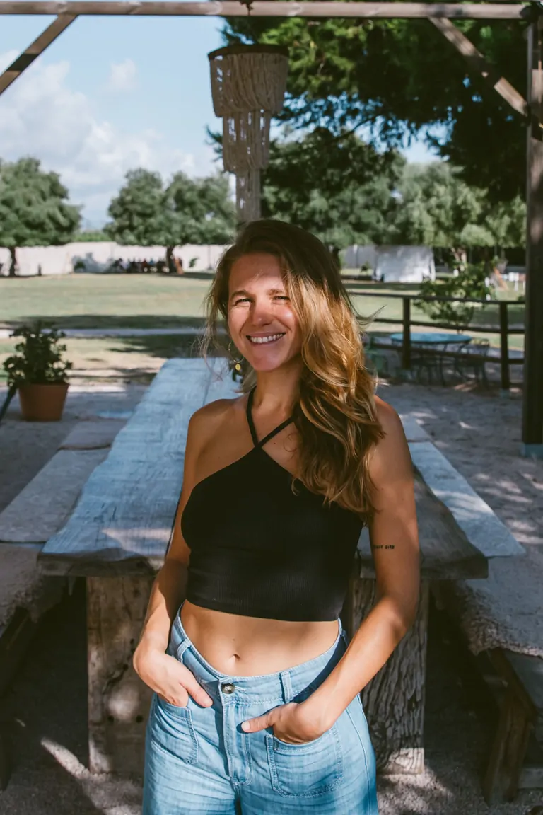 Aina, the Kitachen & Booking Manager at Suniai Oliva, smiles warmly in a sunny outdoor setting by a rustic wooden table.